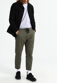 Chaqueta negra abotonada, camiseta gris, pantalones cargo verde oliva con bolsillos, zapatillas negras y calcetines blancos. Conjunto informal con colores contrastantes.