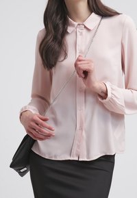 Blusa lunga a maniche lunghe di colore rosa chiaro con colletto, patta con bottoni e polsini elasticizzati. Abbinata a una gonna nera e una borsa piccola.