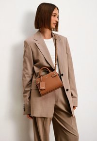 Sac à main en cuir marron avec un design structuré, de courtes poignées et une bandoulière amovible. Il dispose d'une petite étiquette et de subtils accents en matériel.