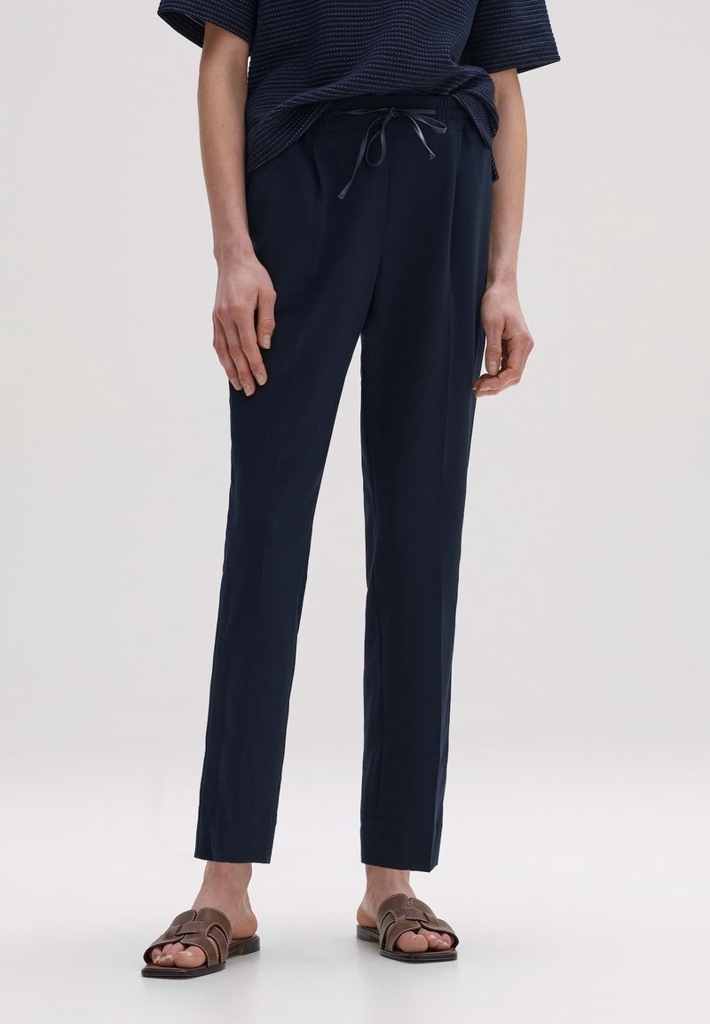 Personne portant un pantalon bleu marine à cordon, associé à des sandales slides foncées et un haut bleu marine texturé à manches courtes, debout sur un fond clair.
