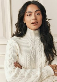 Cream cable-knit sweater with a high collar, featuring textured patterns and ribbed cuffs. Styled with wavy dark hair and gold jewelry accents.