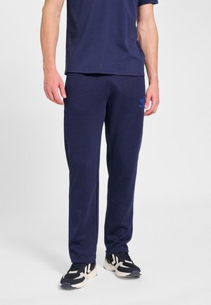 Mand iført marineblå joggingbukser og matchende t-shirt, kombineret med sort-hvide sneakers, stående på en ensfarvet hvid baggrund.