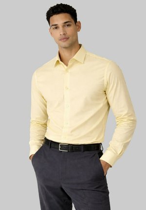 Homme portant une chemise pale jaune à manches longues boutonnée, rentrée dans un pantalon gris foncé, les mains dans les poches, sur un fond gris.