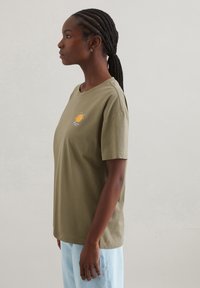 Olivgrünes Baumwoll-T-Shirt mit lockerer Passform, Rundhalsausschnitt und kurzen Ärmeln. Verfügt über ein kleines gesticktes Design auf der Vorderseite.