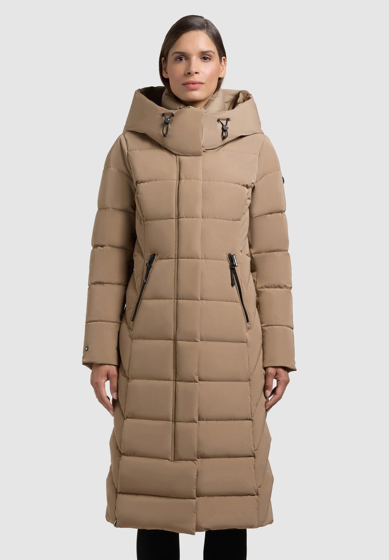 Dlouhý béžový péřový kabát s kapucí, s horizontálním prošívaným vzorem, dvěma předními kapsami na zip a přezkami pro nastavení otvoru u krku.