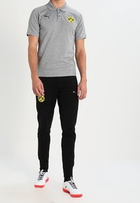 Homme portant un polo gris et un pantalon noir avec des logos jaunes BVB 09, debout avec des crampons blancs et rouges sur un fond blanc.
