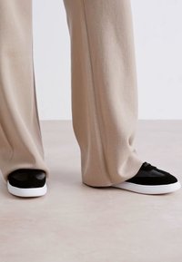 Sneaker bianchi e nere con una tomaia testurizzata, caratterizzate da una punta nera liscia e una suola in gomma bianca, abbinate a pantaloni beige a gamba larga.