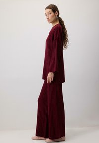 Burgundský úpletový set na domácí nošení, který obsahuje dlouhý rukáv s širokými rukávy a volně střižené pyžamové kalhoty, které ukazují texturovaný žebrovaný vzor.