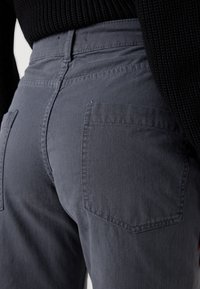 Közelkép egy személyről, aki sötétszürke farmernadrágot és fekete kötött pulóvert visel, a hátsó zseb és a derékpánt varrására összpontosítva.