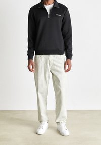 Sudadera negra con cremallera y cuello, con un logotipo blanco. Combinada con pantalones de rayas color crema y zapatillas blancas sobre un suelo beige.