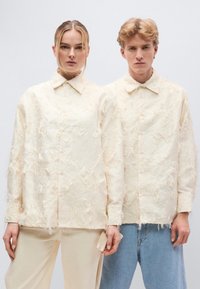 Κρεμ πουκάμ ισα με υφή και σκισμένες λεπτομέρειες, σε συνδυασμό με ανοιχτόχρωμο παντελόνι. Casual εφαρμογή, κλασικό γιακά και διακριτική λάμψη.