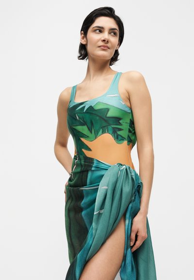 Femme portant une robe à imprimé tropical vert et bleu avec une fente haute, regardant sur le côté sur un fond blanc uni.