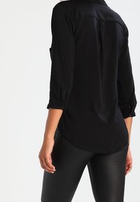Camisetta nera a maniche lunghe con orlo curvo, polsini bottonati e una texture liscia; presenta una cucitura centrale sul retro.