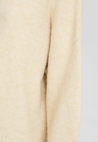 Beiger Strickpullover mit weicher Textur, ausgestattet mit einem Rundhalsausschnitt und einem Design mit überschnittenen Schultern. Rippbündchen und Saum sorgen für Struktur.