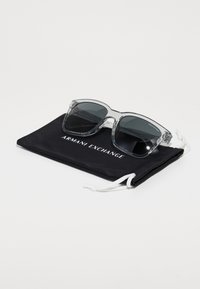 Ochelari de soare gri transparenți cu lentile închise, pe un pachet din material textil negru, având imprimat în alb "ARMANI EXCHANGE".