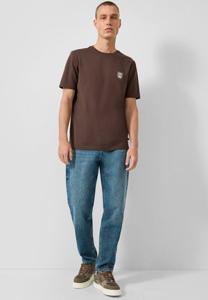 Mężczyzna ubrany w brązowy krótki t-shirt z małym logo, niebieskie dżinsy i brązowe trampki, stojący na jednolitym jasnoszarym tle.