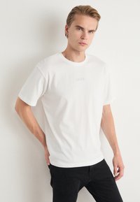 Hvit bomull T-skjorte med rund hals og korte ermer, med en subtil preget "BOSS" logo på brystet. Kombinert med svarte bukser.