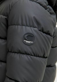 Černá péřová bunda s prošívanými sekcemi, hladkým povrchem a kulatou látkovou nášivkou s názvem značky na rukávu.