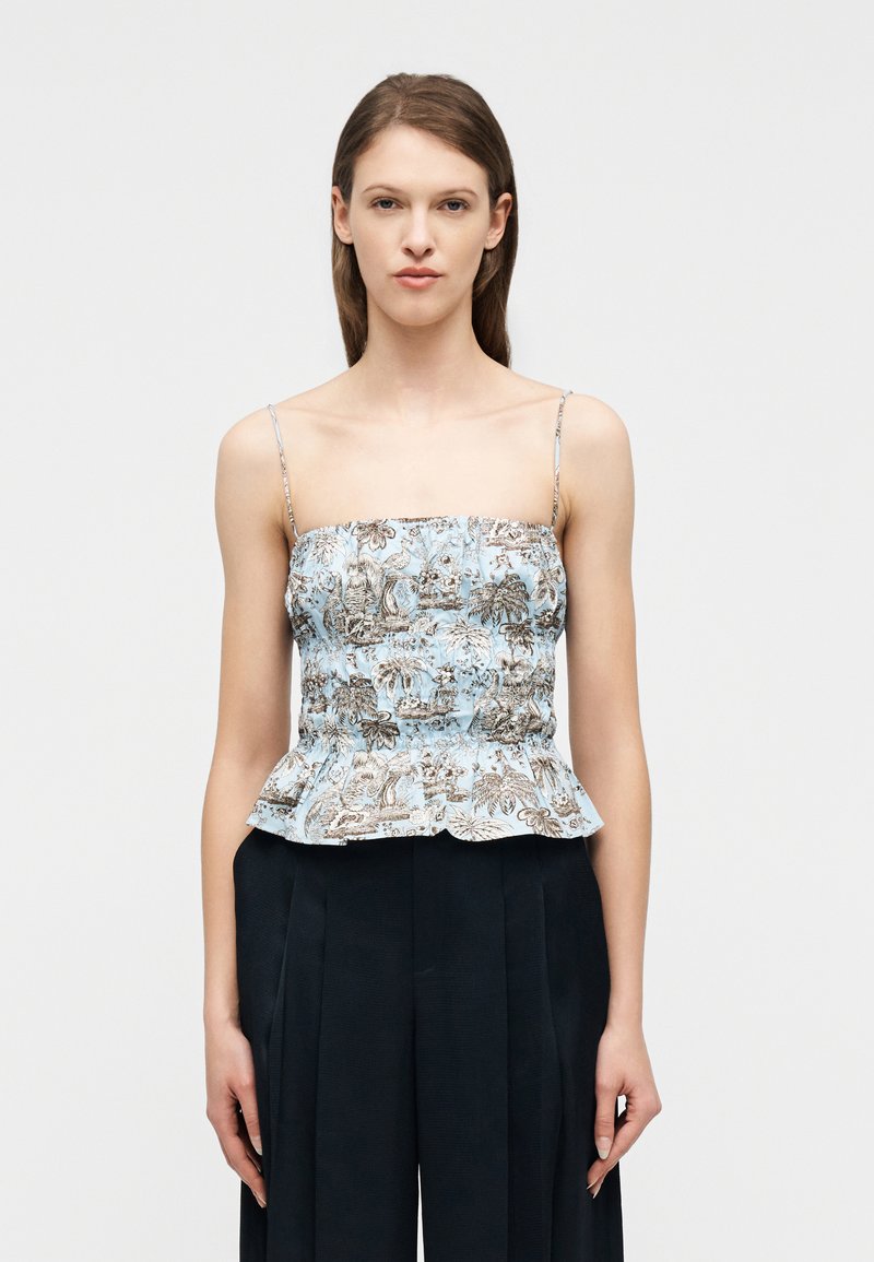 Γυναίκα με αμάνικο, ανοιχτό μπλε τοπ πεπλούμ με λουλούδια και μαύρο ψηλοκάβαλο παντελόνι με φαρδιά πόδια, σε απλό λευκό φόντο.