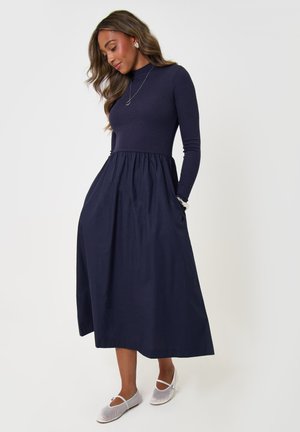Woman in a long navy dress with a fitted top and full skirt, wearing white flat shoes, standing against a plain light background.