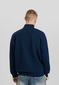 Sudadera azul marino con cuello alto y puños acanalados, fabricada en un tejido suave; presenta un corte holgado y un diseño minimalista.