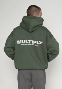 Grøn hættetrøje med en stor hvid "MULTIPLY" logo på ryggen. Har en rummelig hætte og løs pasform. Fremstillet af blødt, struktureret stof.