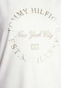 Camiseta blanca de algodón con texto metálico plateado que dice "TOMMY HILFIGER", "Nueva York" y "EST. MCMXLXX". Logo en el centro.