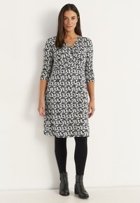 Kvinna står mot en vit vägg klädd i en svart och vit blommig klänning, svarta leggings och svarta ankelstövlar.