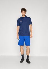 Camisa polo azul-marinho com colarinho e três botões, combinada com calções de desporto azul brilhante com detalhes de logo, usados com sapatos pretos.