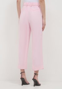 Donna che indossa pantaloni cropped rosa chiaro con orli risvolti e sandali con tacco neri, in piedi su uno sfondo bianco semplice.