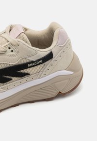 Shoe atlético em bege com parte superior em malha e camurça, detalhes do logótipo em preto, colarinho almofadado e sola de borracha texturizada com acentos em bege.