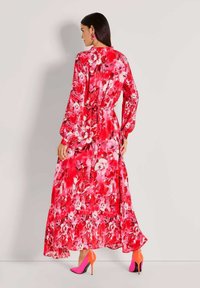 Blumiges Maxikleid in lebendigen Rottönen mit langen Ärmeln, gebundener Taille und fließendem Saum. Kombiniert mit leuchtend pinken Absätzen.