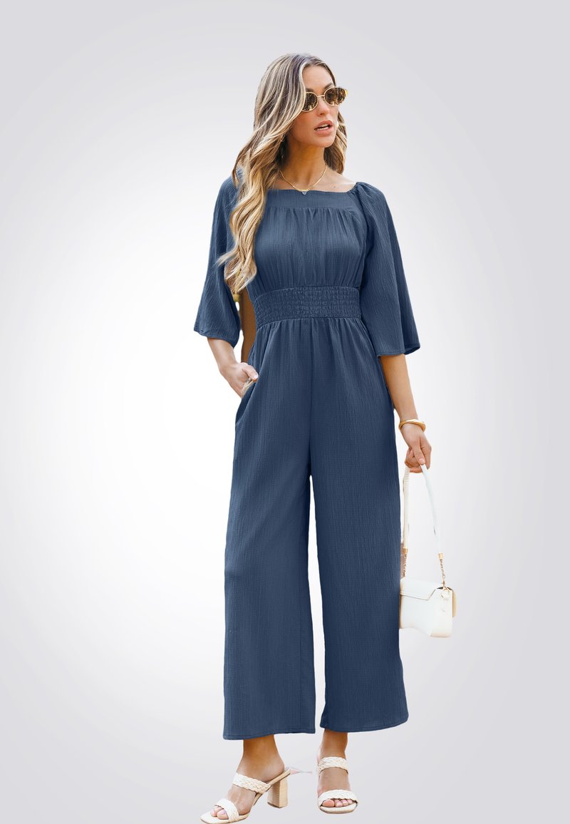 Navyblauwe jumpsuit met een vierkante halslijn, korte mouwen, een geraffineerde taille, wijde benen en zijzakken. Draag het met witte haksandalen.