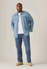 Svetlo modra denim srajca z detajli žepov, pod njo bela majica, v kombinaciji s svetlo modrimi kavbojkami in bež športnimi čevlji.