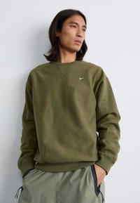 Mlad moški z dolgimi lasmi nosi olivno zeleno majico s kapuco z manjšim belim logotipom Nike, ki stoji pred enobarvno ozadjem.