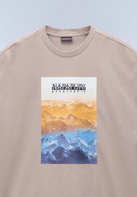 Béžové bavlněné tričko s grafikou horské krajiny v odstínech oranžové a modré, s textem "NAPAPIJRI geographic" nad obrázkem.