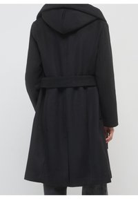 Long manteau noir avec capuche et ceinture, vu de dos sur une personne debout devant un fond clair uni.