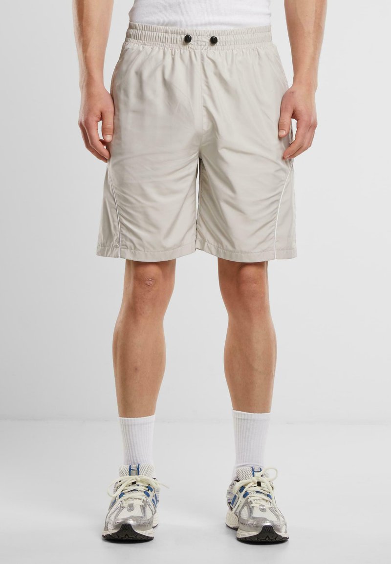 Beige Sport Shorts mit elastischem Bund, zwei schwarzen Knöpfen, Seitentaschen und weißen Nähakzenten. Hergestellt aus leichtem Material.