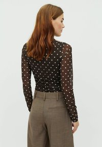 Long-sleeve, sheer black top with white polka dots, fitted at the waist, paired with light brown, tailored trousers.