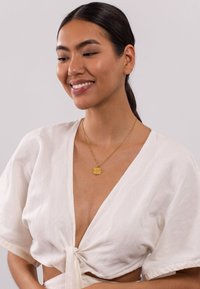 Goldener Anhänger an einer feinen Kette, kombiniert mit einer weißen Leinenbluse mit tiefem V-Ausschnitt und Taillenschnürung.