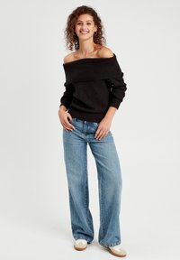 Sort off-the-shoulder sweater med ribbet halsudskæring, parret med lyseblå vidde ben jeans. Modellen står med hænderne på hoften og smiler.