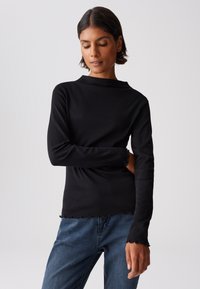 Schwarzes Langarmoberteil mit rundem Ausschnitt und gerafftem Saum. Hergestellt aus weichem Stoff mit taillierter Passform und strukturierten Ärmeln. Kombiniert mit blauen Jeans.