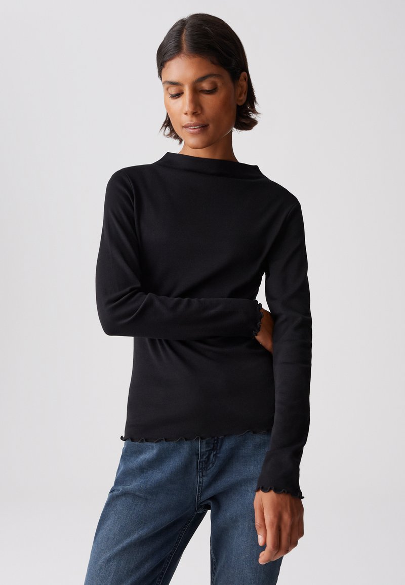 Schwarzes Langarmoberteil mit rundem Ausschnitt und gerafftem Saum. Hergestellt aus weichem Stoff mit taillierter Passform und strukturierten Ärmeln. Kombiniert mit blauen Jeans.