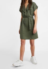Abito verde oliva con cintura, maniche corte, tasche sul petto e vestibilità rilassata, abbinato a sneakers bianche. Tessuto liscio e leggero.