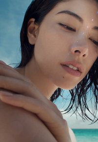 Nahaufnahme eines Porträts einer Person mit nassen, dunkelbraunen Haaren, subtilen Sommersprossen und glatter Haut, umrahmt von einem blauen Himmel und einem Ozeanhorizont.