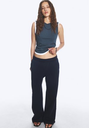Junge Frau mit langen, welligen Haaren trägt ärmelloses blaues Crop-Top, lockere schwarze Hose, Sandalen und Schmuck vor hellem einfarbigem Hintergrund.