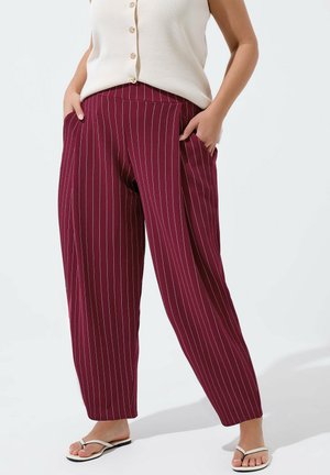 Kvinde iført højttaljede bordeaux pinstripe bukser med vide ben, kombineret med en hvid ærmeløs knapoplukningsbluse og hvide flip-flops.