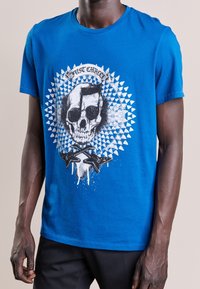 Blå bomullströja med en dödskallegrafik som har musikaliska inslag och ett cirkulärt mönster. Kortsleeves och design med rund hals.