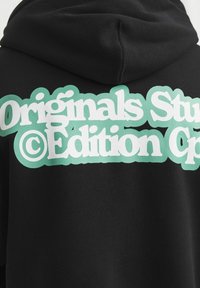 Sort hættetrøje med hvid og grøn tekst, der lyder "Originals Studio Edition" på øverste del af ryggen under hætten.
