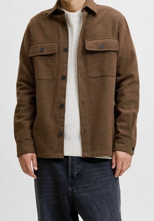 Mann trägt braune Hemdjacke über weißem Strickpullover und dunklen Jeans, steht vor weißem Hintergrund.